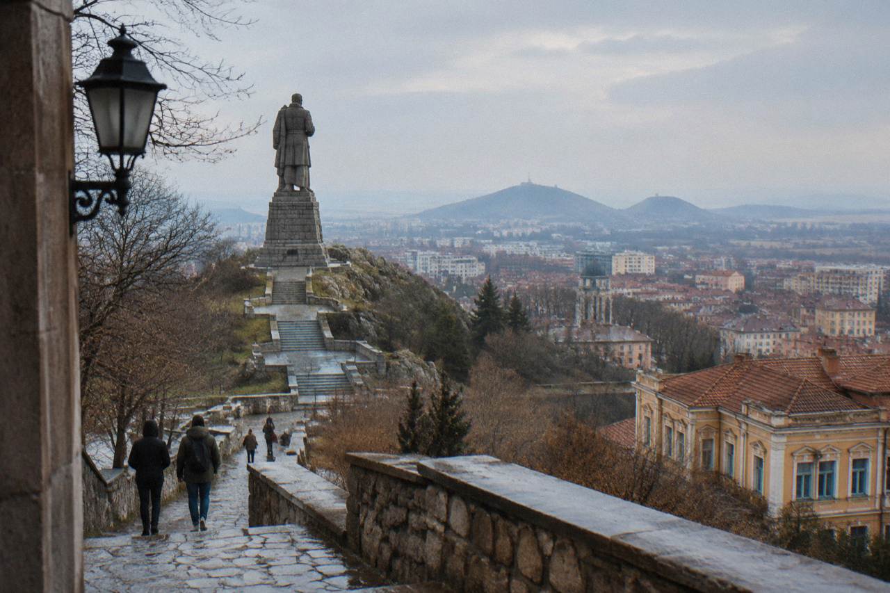 A tall stone statue of a soldier standing on a pedestal above a green park hill above a city skyline