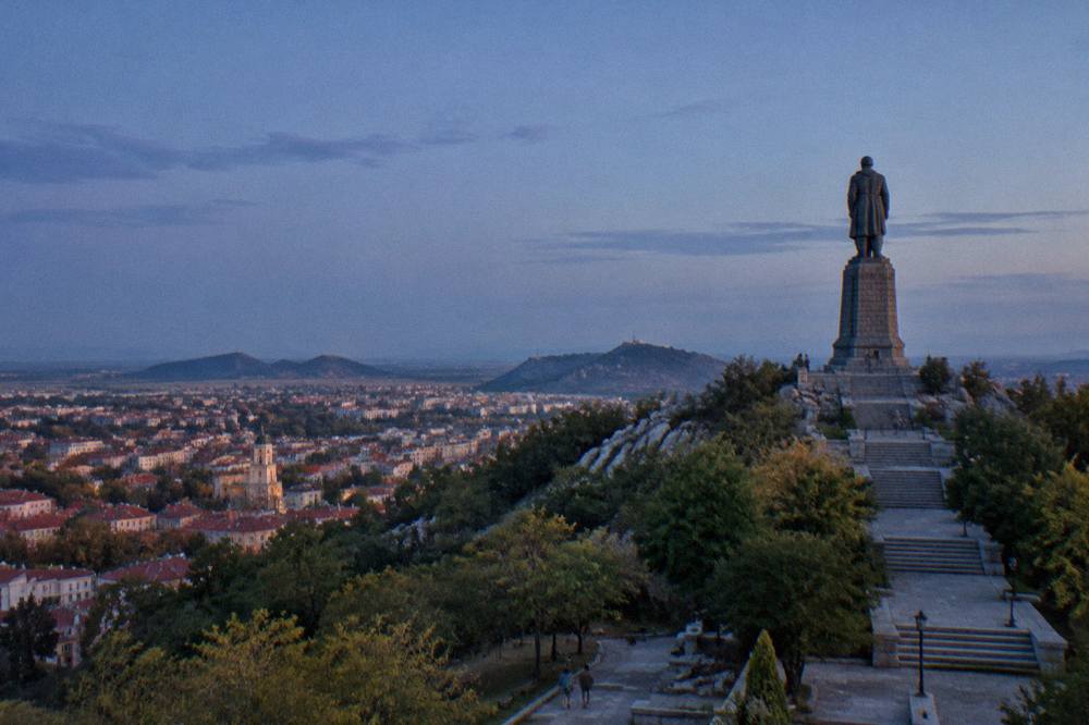 A wide view of a hilltop park with a tall soldier statue at the summit and city rooftops spreading below