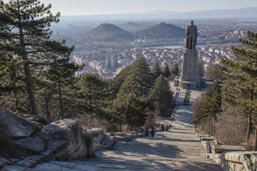 Stone steps climbing through a pine park toward a hilltop statue with city buildings visible below
