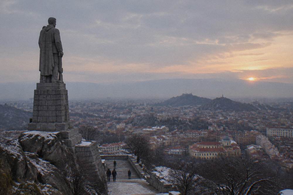 A hilltop statue silhouette against an evening sky with a city of red rooftops and distant mountains below