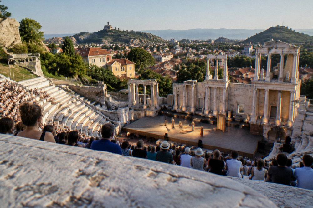 An audience seated in tiered ancient stone seats facing an illuminated stage