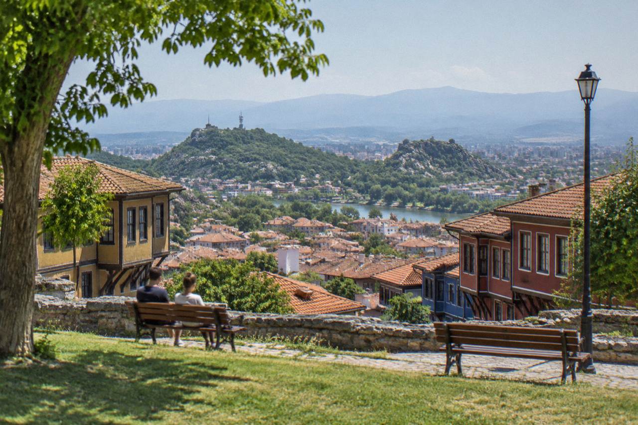 A grassy hilltop park in Plovdiv with benches and a wide view over red rooftops toward distant mountains