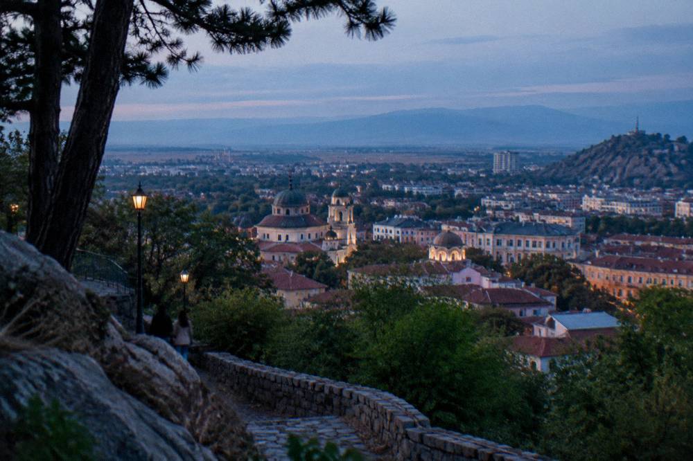 An evening panorama from a hilltop over a city with church domes and distant mountains