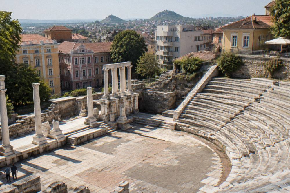 Ancient stone foundations and column bases of a Roman forum square in a Bulgarian city, with modern buildings behind