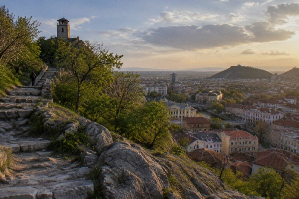 An evening cityscape seen from a hilltop path with warm light on distant rooftops