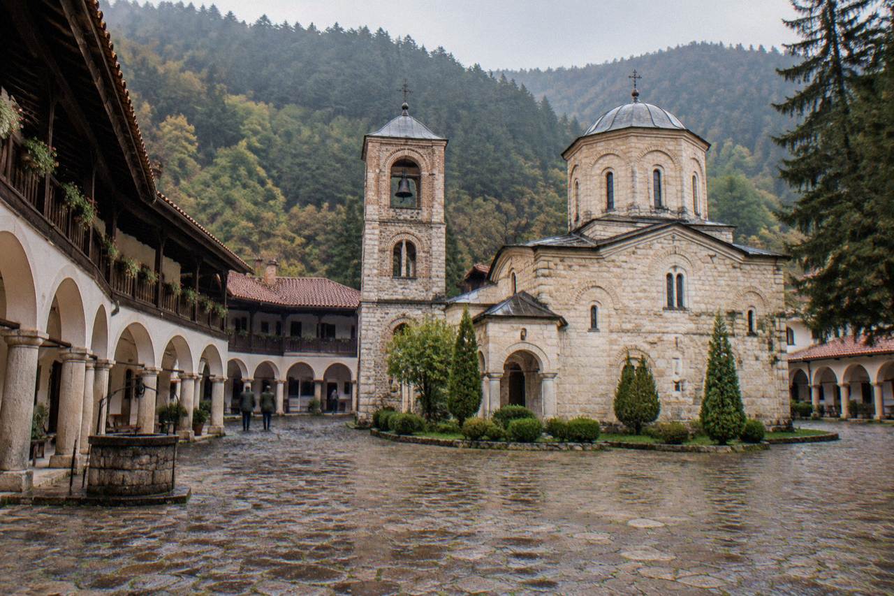 A stone monastery complex with arched cloisters and a church tower set against a green valley