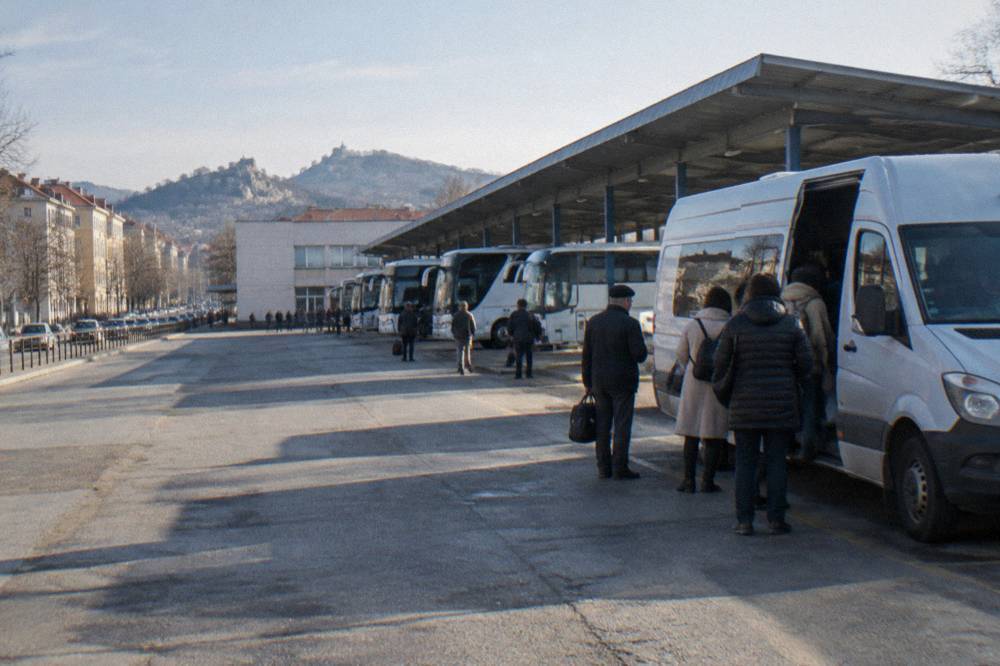 A white minibus parked at a small bus station with passengers waiting to board