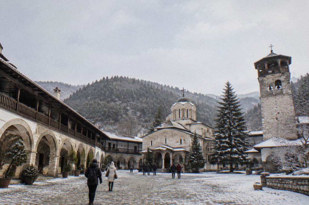 A monastery courtyard with stone arches, visitors walking and a bell tower above
