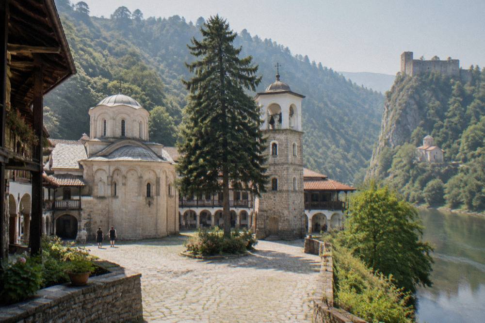 A stone fortress on a cliff top above a river valley with a small church at its base