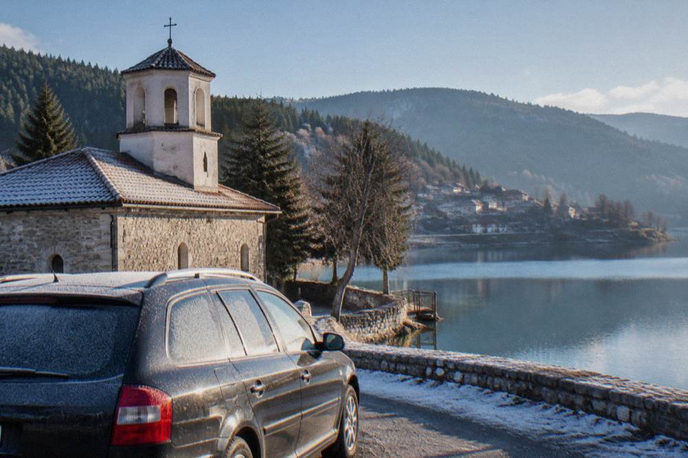 A parked car beside a lakeside road with forested slopes behind