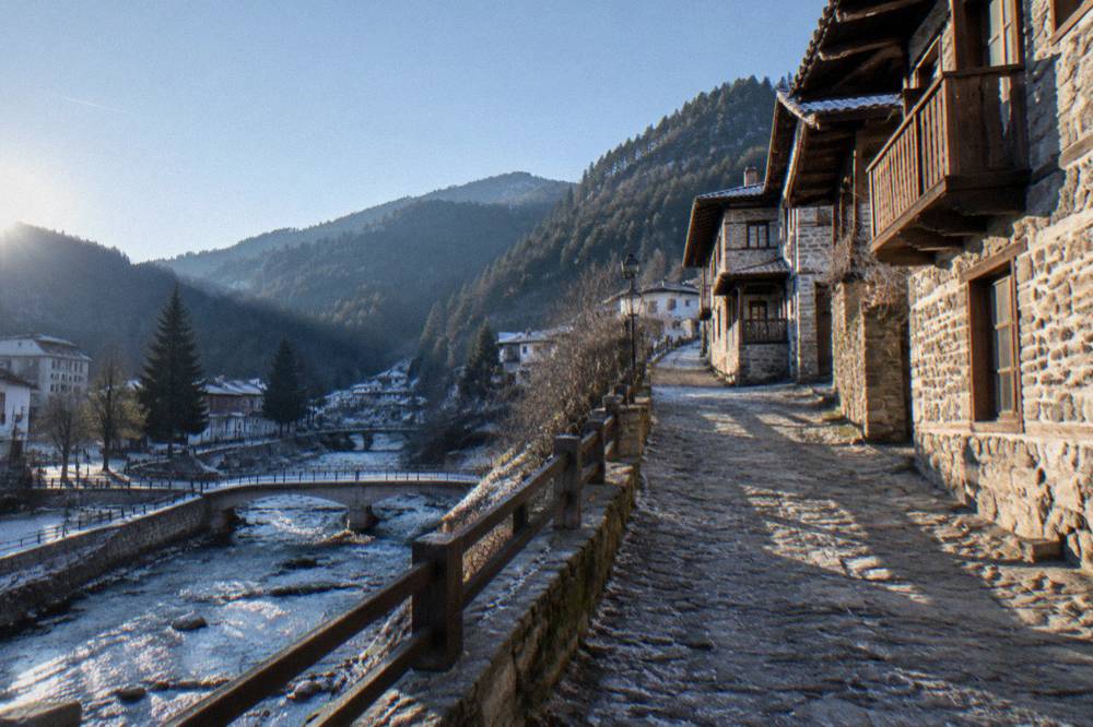 A stone-built Rhodope village street with wooden balconied houses and hills behind