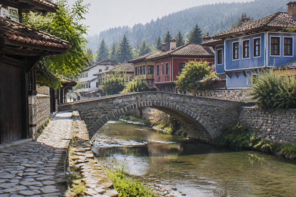 Stone bridge over a small river with coloured revival-era houses behind it