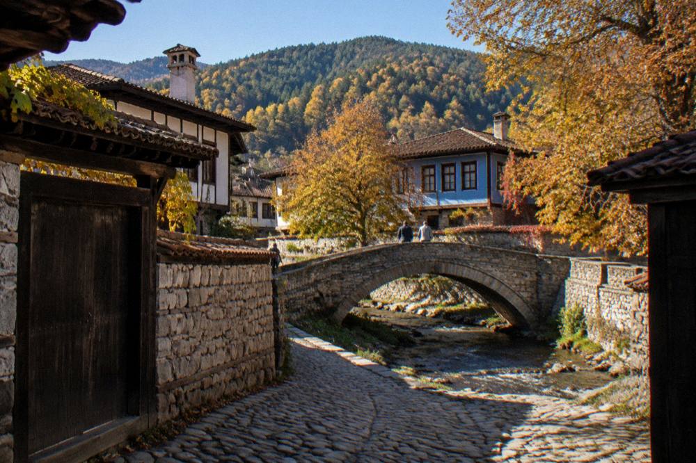 Autumn trees over a stone bridge and a blue sky above mountain roofs