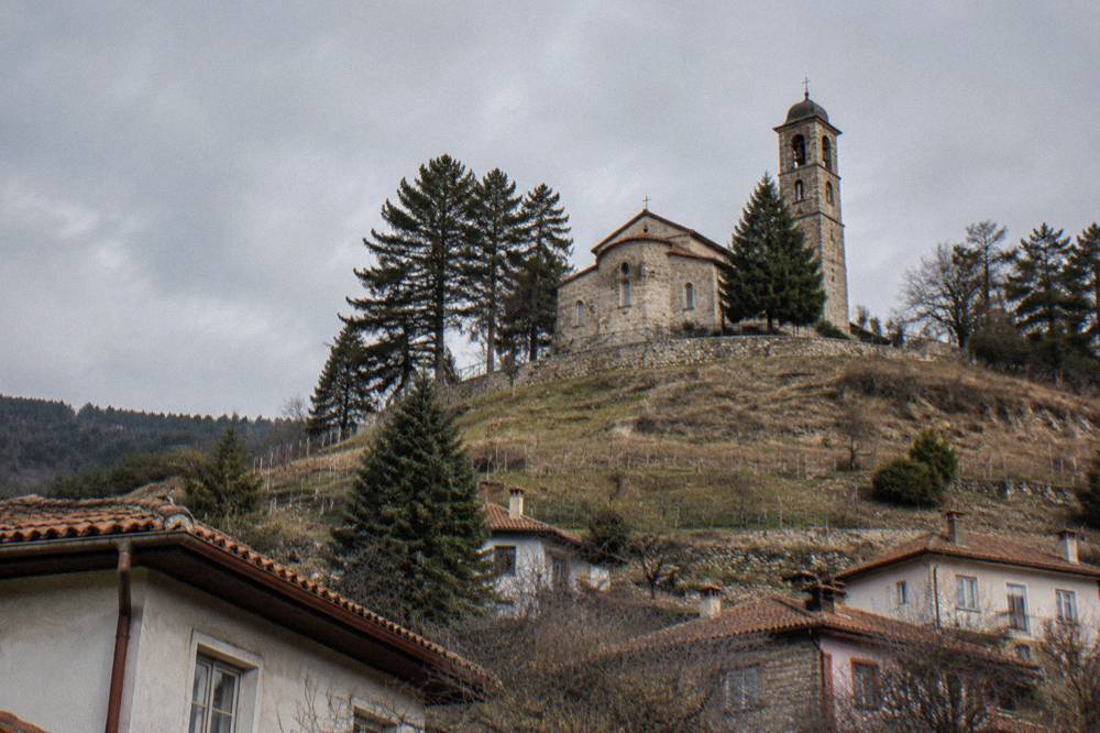 A stone memorial church with a bell tower standing on a grassy hill above a small town
