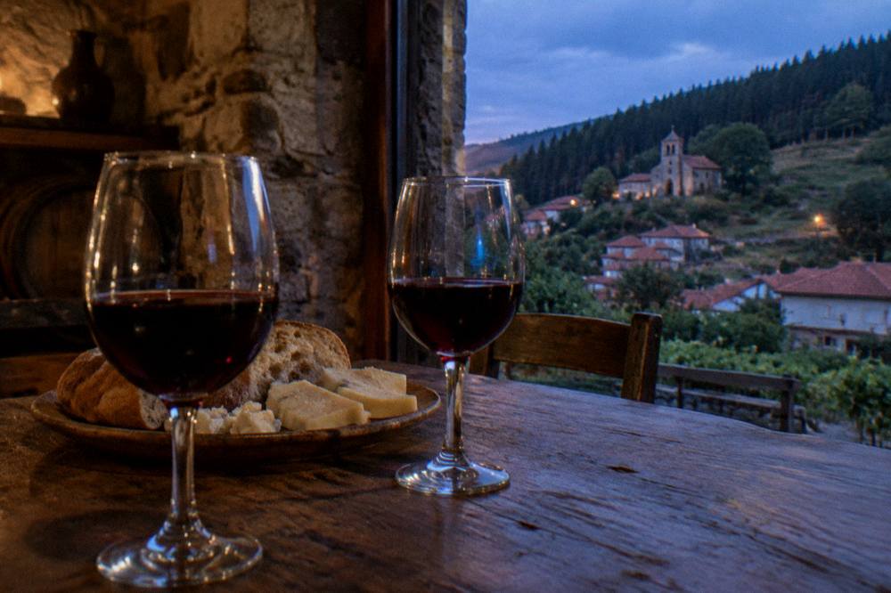 Two glasses of red wine on a wooden table beside a plate of cheese and bread in a rustic cellar