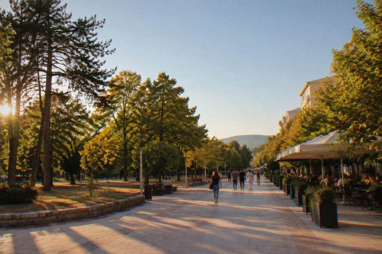 A wide pedestrian boulevard in a southern Bulgarian city lined with trees and cafe tables under a clear sky