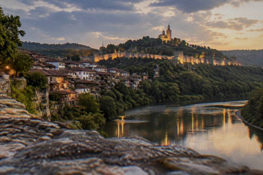 An illuminated fortress on a hill above a town at dusk with lights reflecting in a river