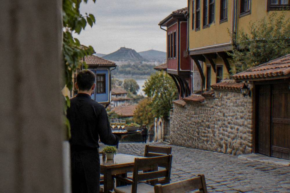 A waiter carrying a tray with rakia glasses and a salad to an outdoor table in Plovdiv