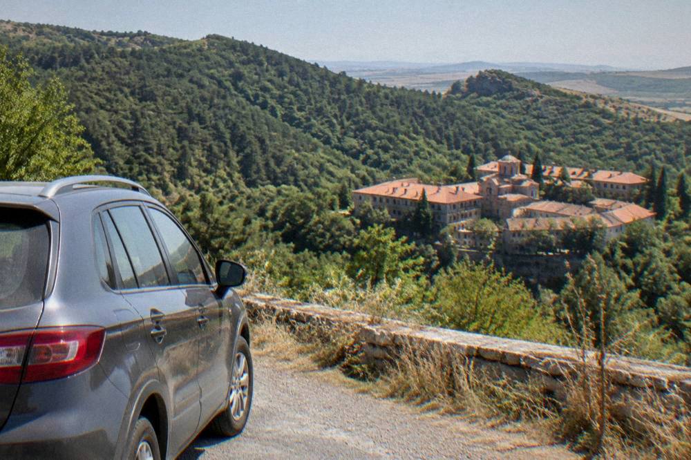 A car parked on a mountain road overlooking a monastery complex in green hills