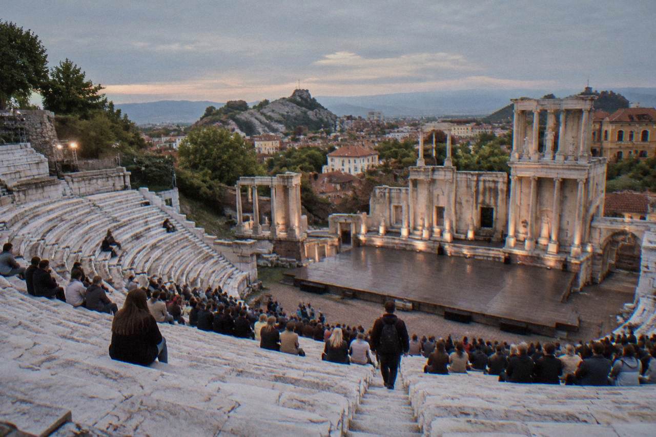 An open-air ancient theatre with marble tiered seats filling with an evening audience