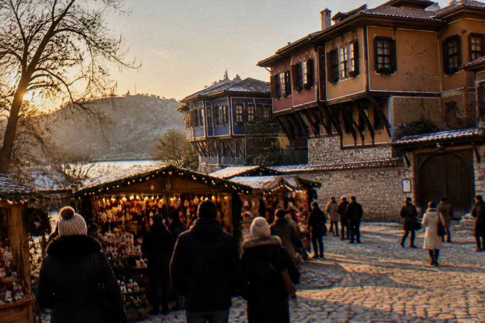 A Christmas market stall with warm lights and a crowd in coats on a Plovdiv square at dusk
