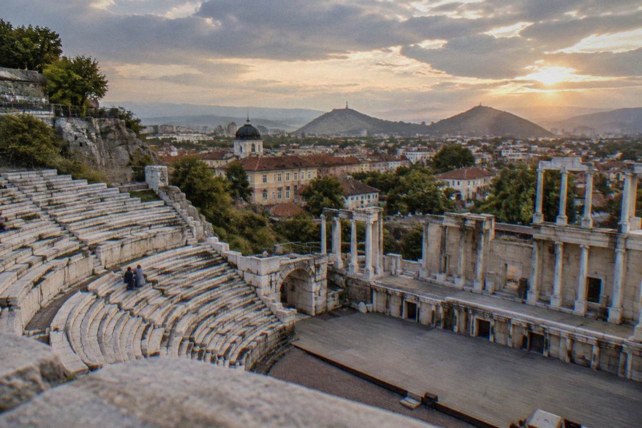 An ancient Roman theatre with marble seat rows rising above a modern city skyline at dusk