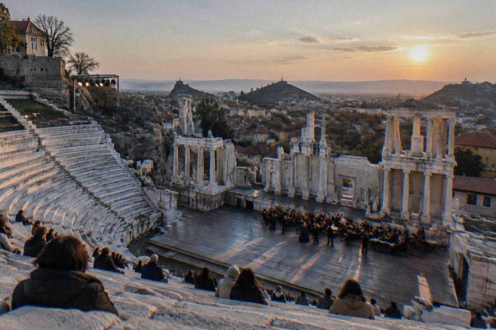 An open-air opera performance underway in an ancient stone theatre under an evening sky