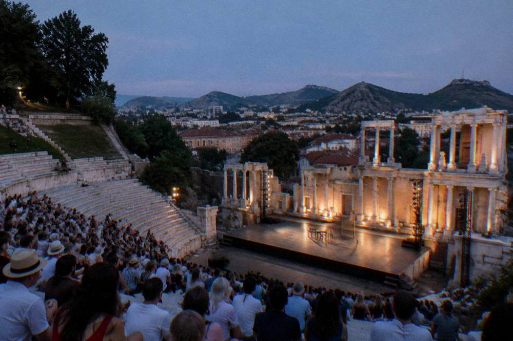 A summer evening crowd seated on stone tiers of an ancient theatre watching a lit stage