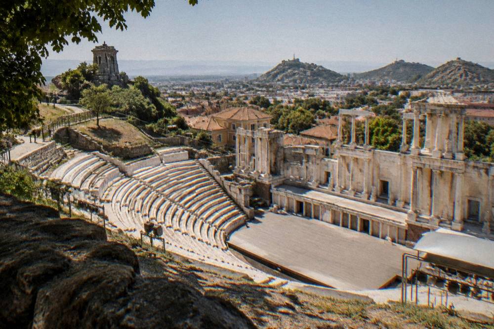 A sunlit hillside path above a city leading toward an ancient stone monument