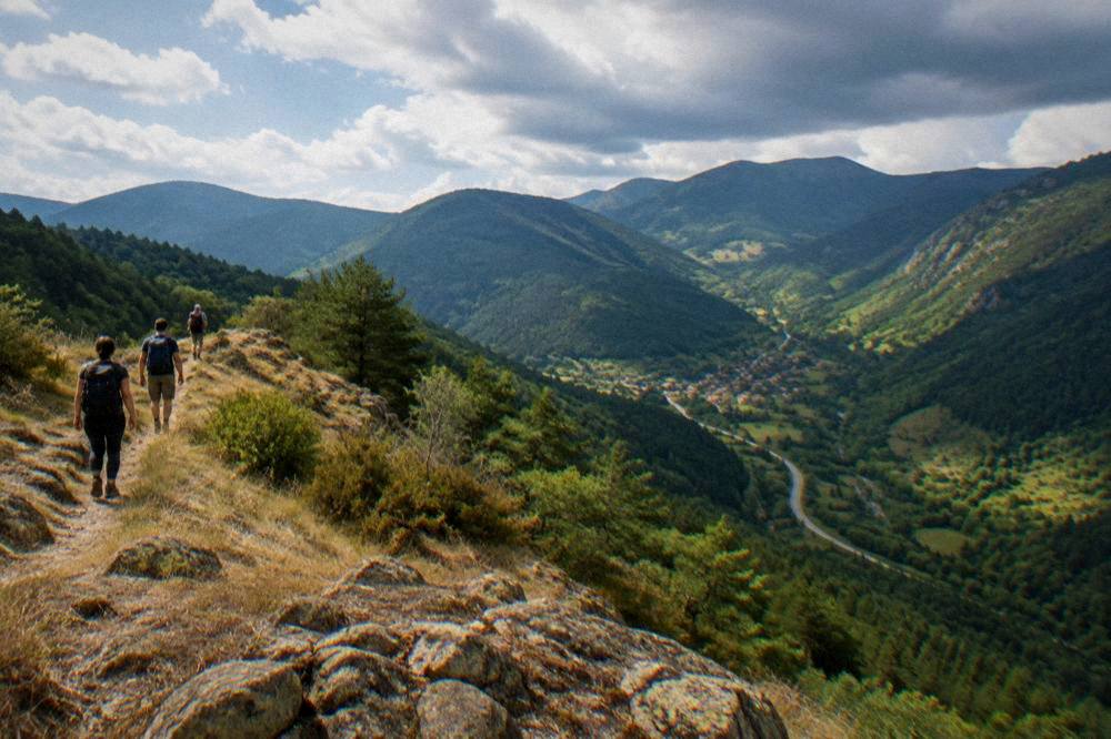 Hikers on a ridge path above a green Rhodope valley with distant village rooftops