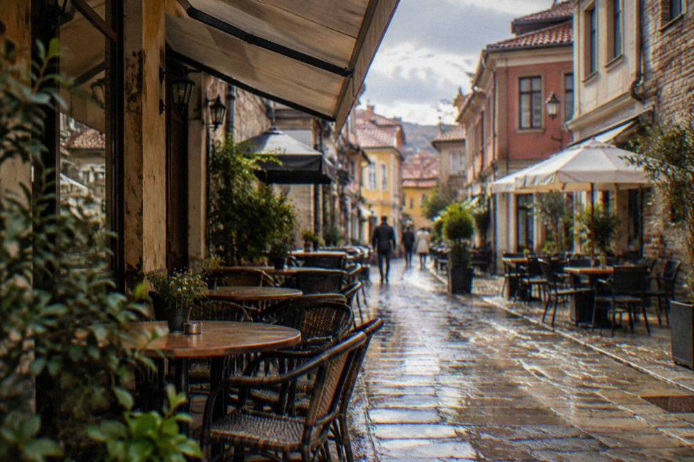 A narrow street in the Kapana district of Plovdiv, cafe tables under awnings, wet cobbles reflecting window light