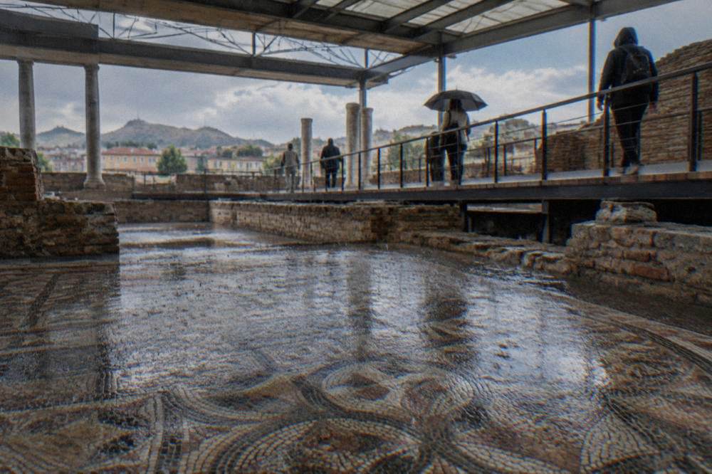 Rain falling on a mosaic floor inside the Bishop's Basilica of Philippopolis, visitors walking a raised walkway