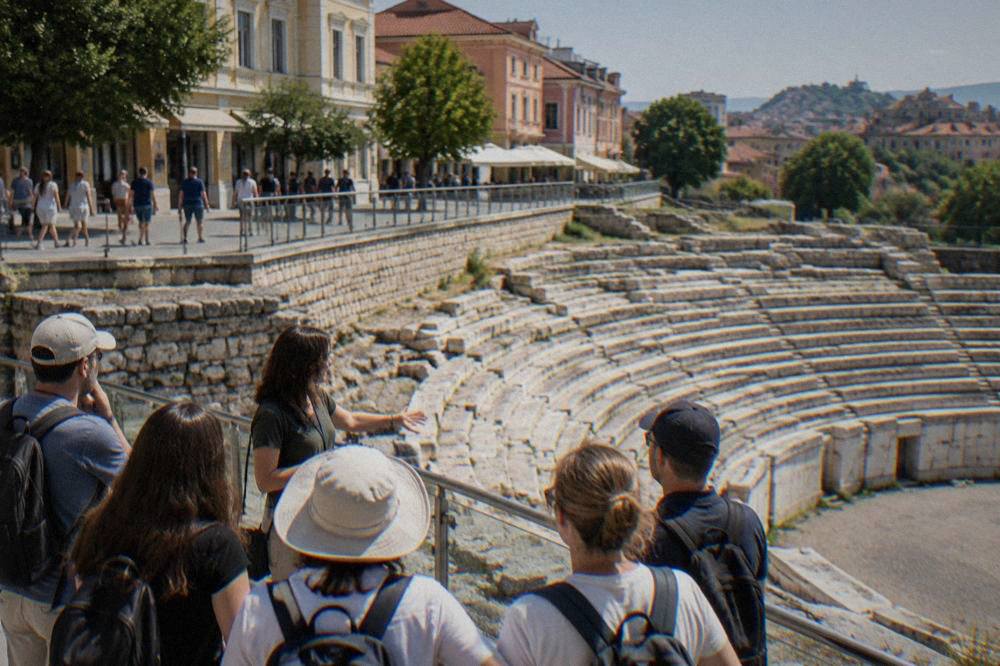 Walkers gathered around a guide beside the exposed stone seats of the Roman Stadium in Plovdiv