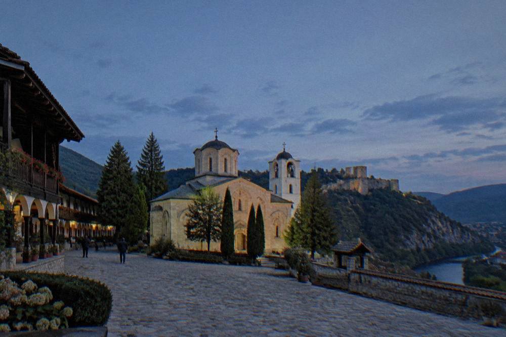 A medieval stone fortress wall on a rocky ridge above a river valley and a small town.